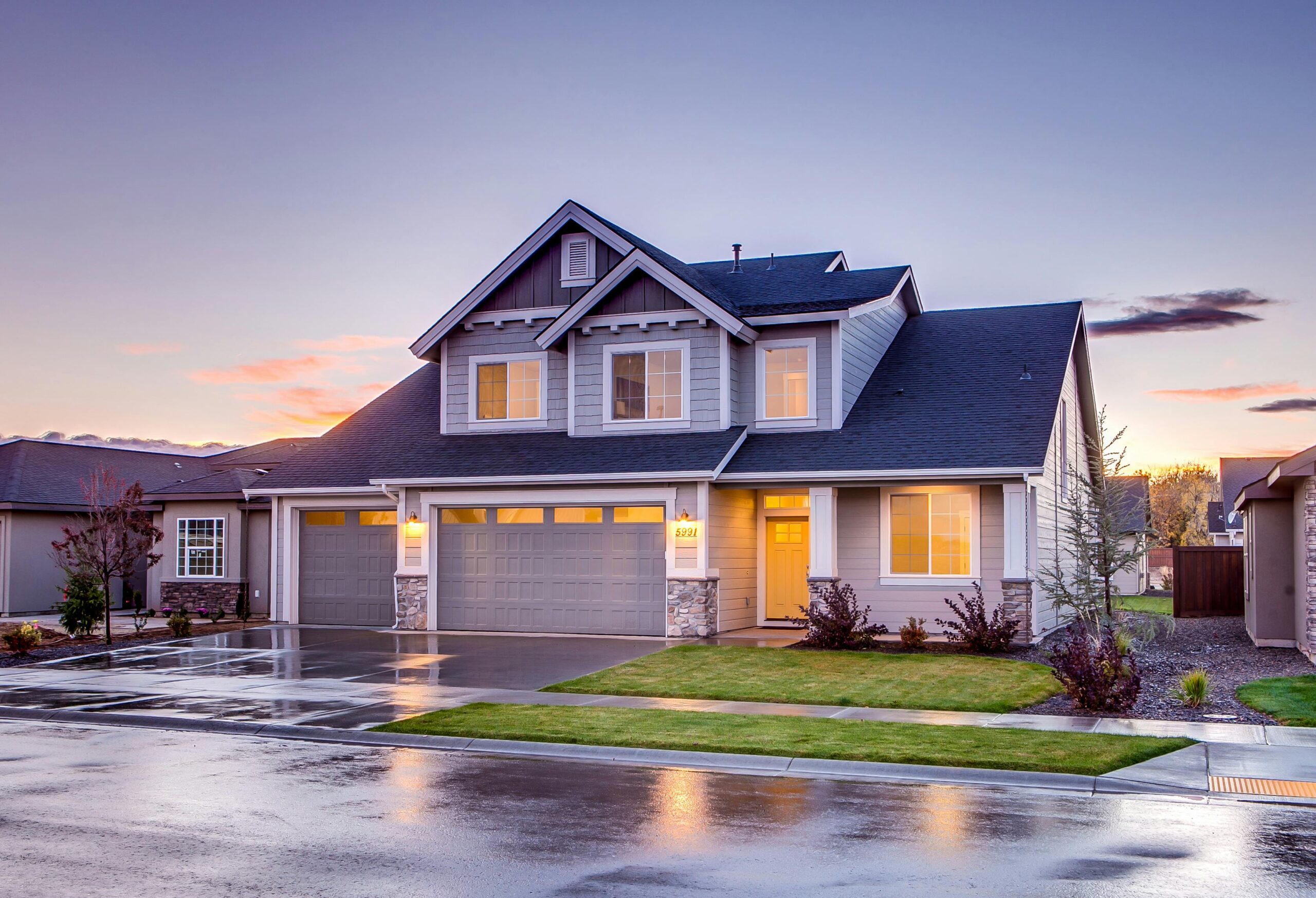 Modern two-story suburban home with three-car garage at sunset, lights on, wet driveway reflecting the sky